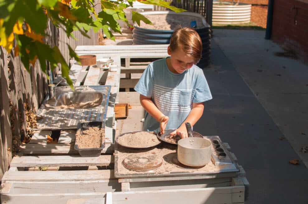 Child plays in backyard in a mud kitchen with pots and pans full of dirt