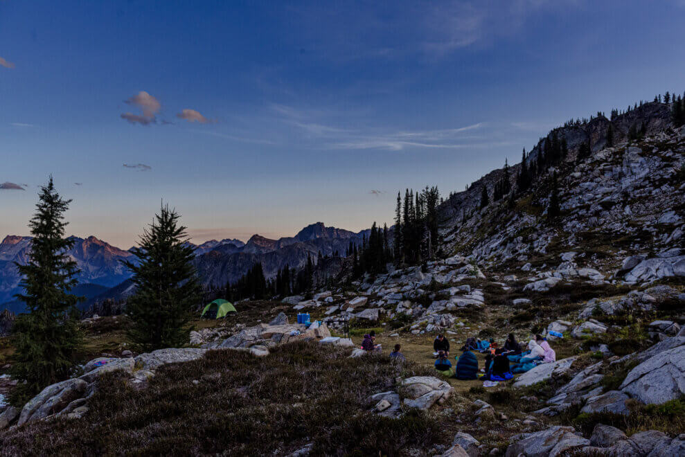 Group of girls sit in a circle in the alpine as the sun sets. A tent sits behind them.