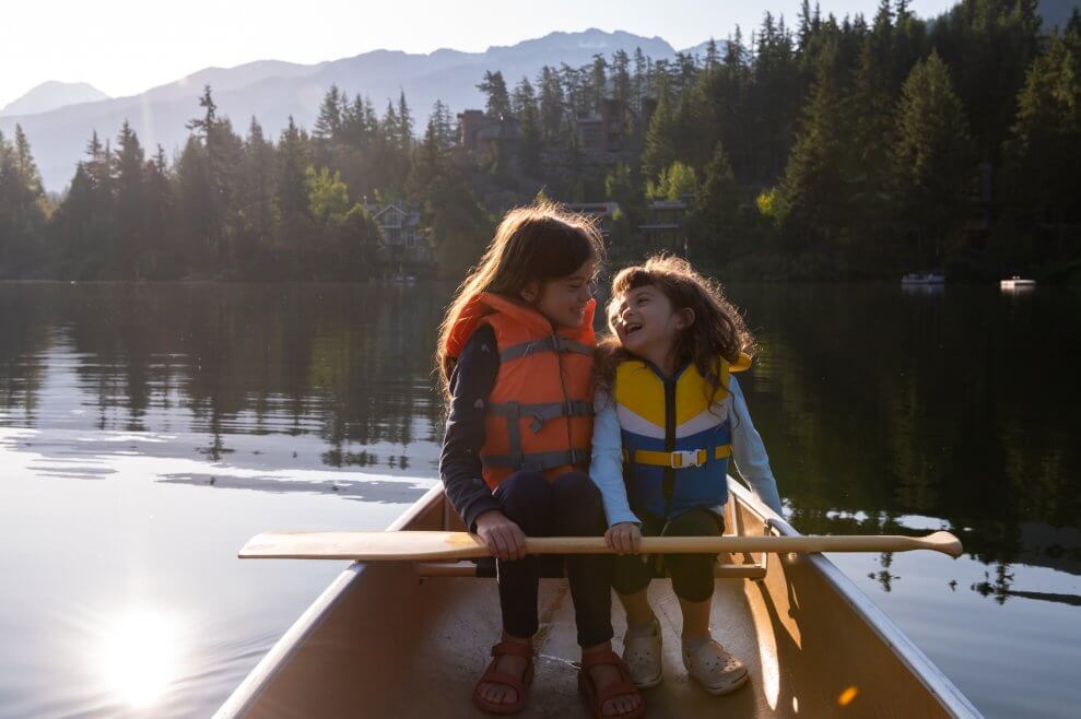 Two sisters sit side by side in a canoe, wearing life jackets and laughing. Canoeing is a great camping activity to do with kids.