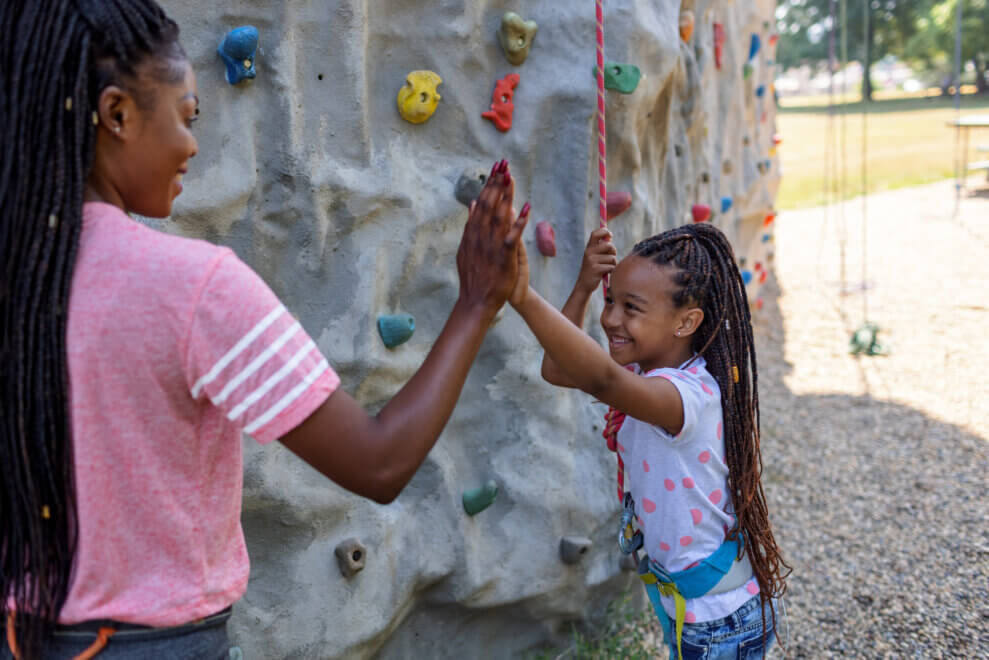 Mom and her daughter high-five in front an outdoor rock-climbing wall. The daughter wears a harness and holds onto a rope with her other hand.