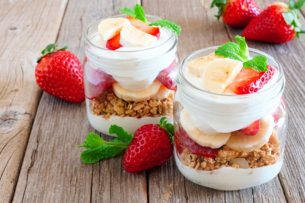 Two jars full of yogurt, granola, strawberries, and bananas sit on a wooden tabletop.