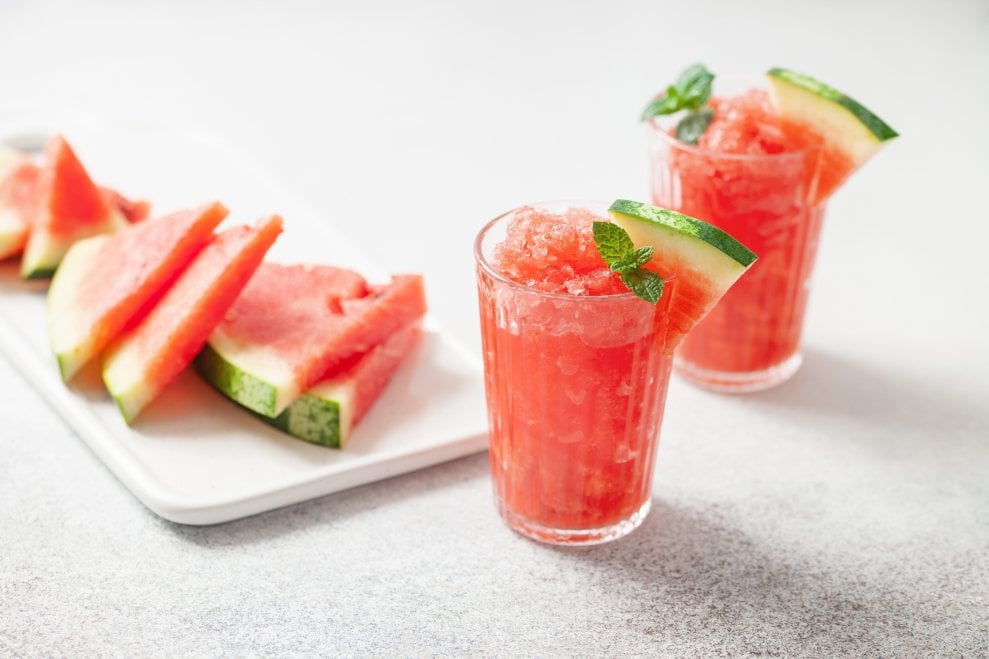 Two glasses of watermelon slush garnished with mint. A cutting board with watermelon slices on it is in the background.