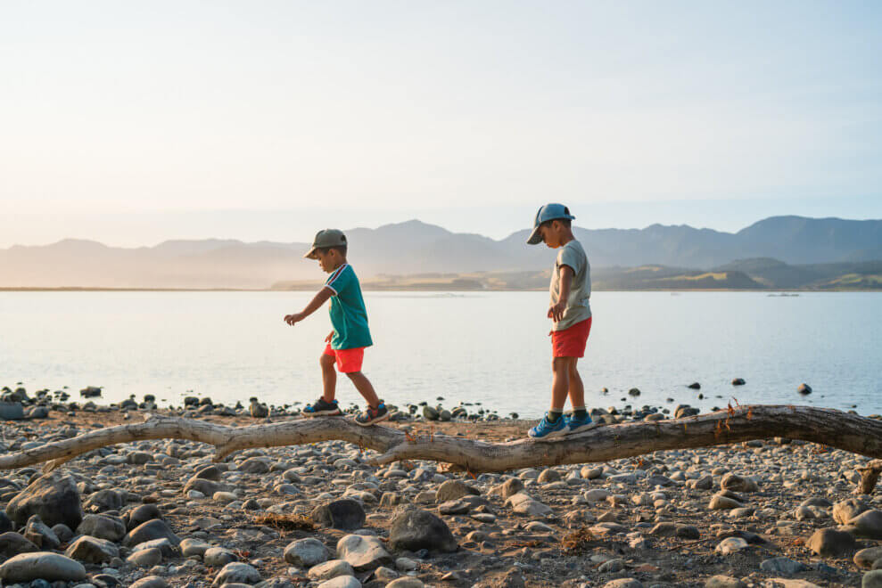 Two young brothers balance as they walk across a log on a beach.