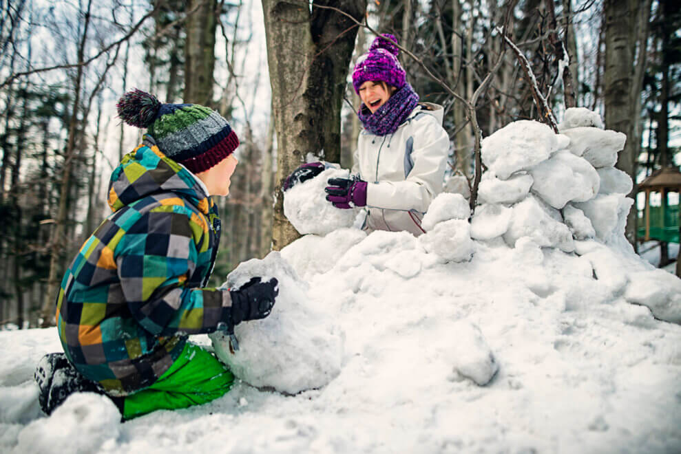 A brother and sister roll snowballs and make a snow fort in the woods.
