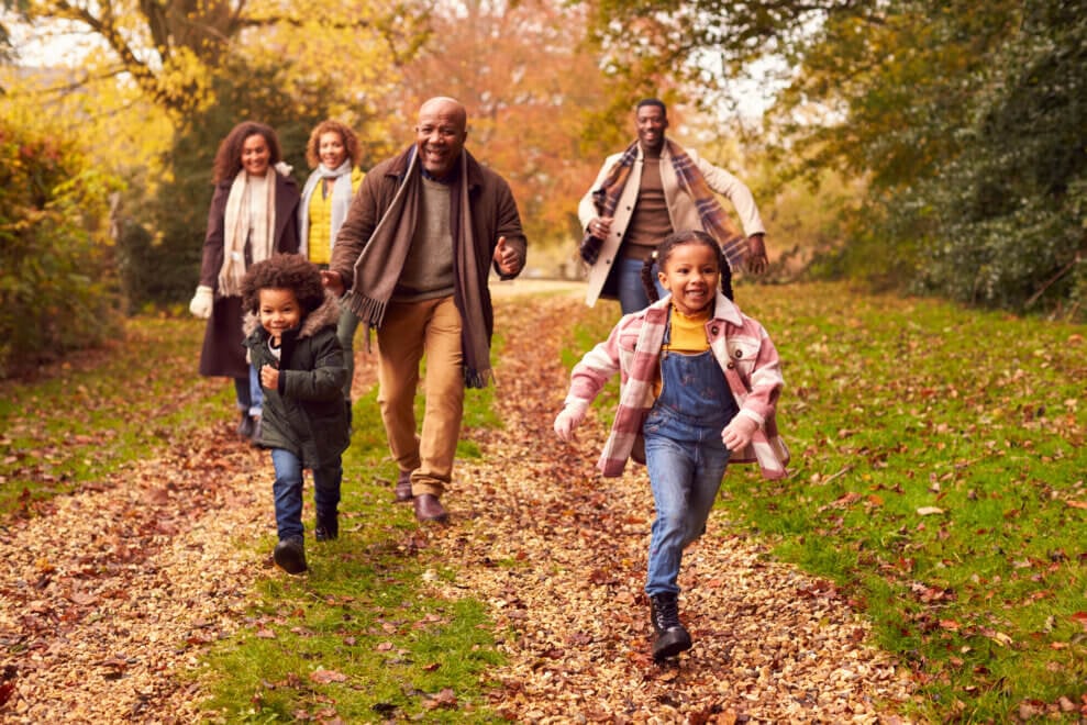 A family walks along a trail in the fall. The grandfather runs after the two young children, laughing, while the two mothers and an uncle walk behind, smiling.