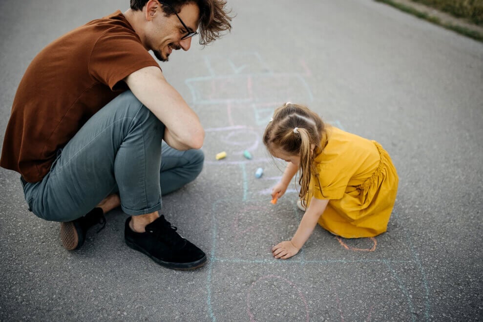 A father and his daughter draw a hopscotch board with chalk on the pavement.