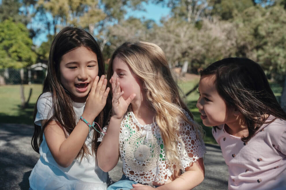 Three girls sit in a row playing Broken Telephone at the park. One girl whispers into the ear of her friend on her right.