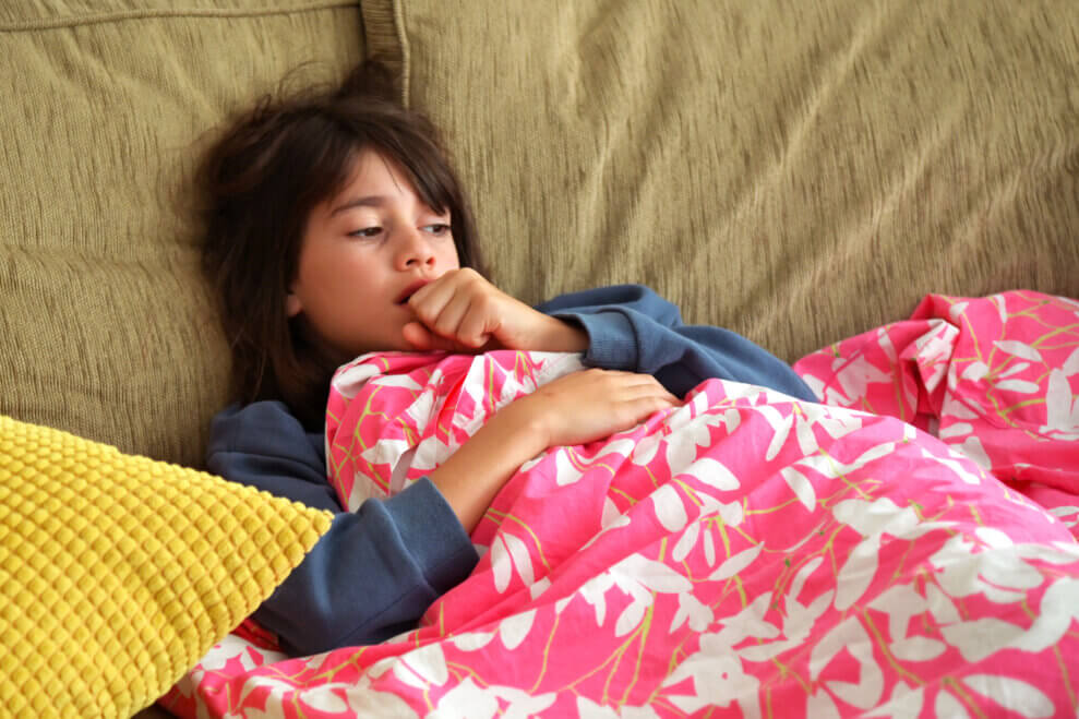 A girl lies on the couch wrapped in a blanket as she coughs