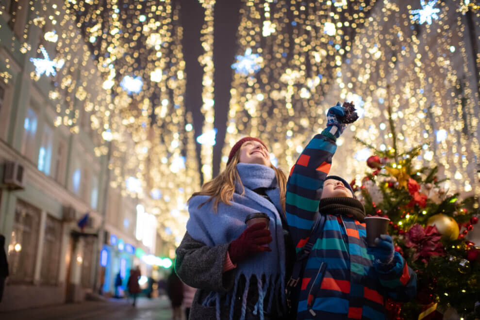 A boy and his mother walk through a street lit with overhead Christmas lights.