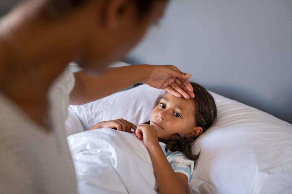 Mother rests a hand on her daughter's forehead as the daughter lies in bed