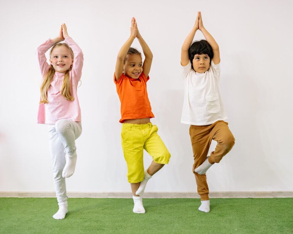 Two girls and a boy stand next to each other in tree pose, with their knee on their lower leg and their hands touching and over their heads.