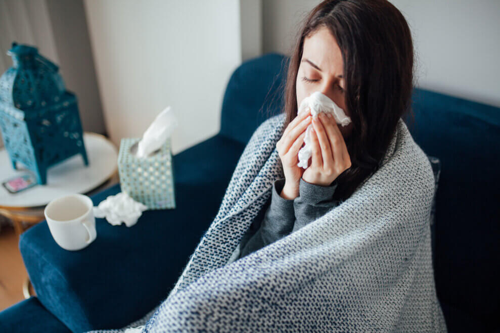 Woman sits on the couch wrapped in a blanket and blows her nose
