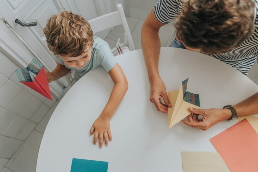 A boy and his father sit at a table and fold paper airplanes.