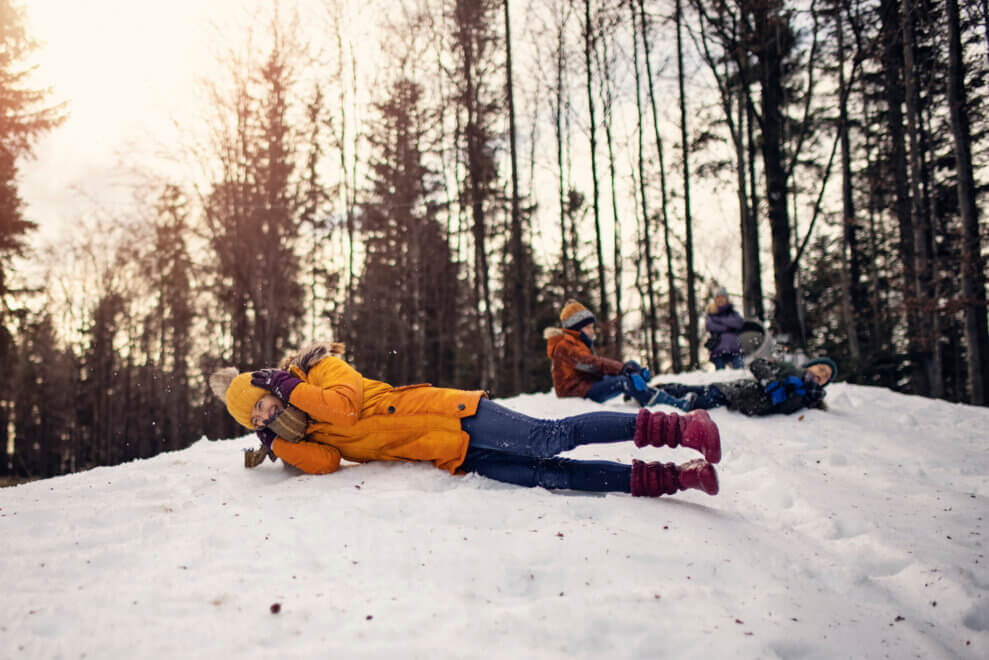 A mom and her three kids take turns rolling down a snowy hill in the winter.