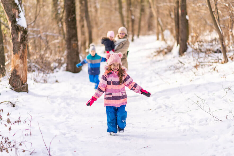 A young girl dressed in a snowsuit runs through the snow in the woods. Her brother and her mother, carrying her toddler sibling, walk behind her.