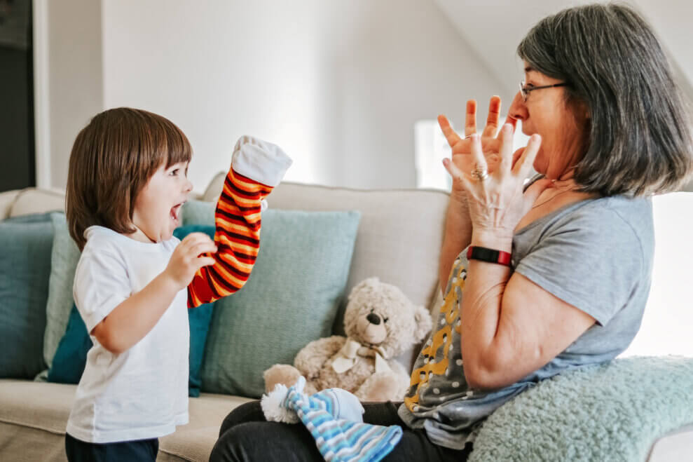 A toddler tries to surprise her grandmother with a sock puppet while the grandmother makes an expression of mock fright. 