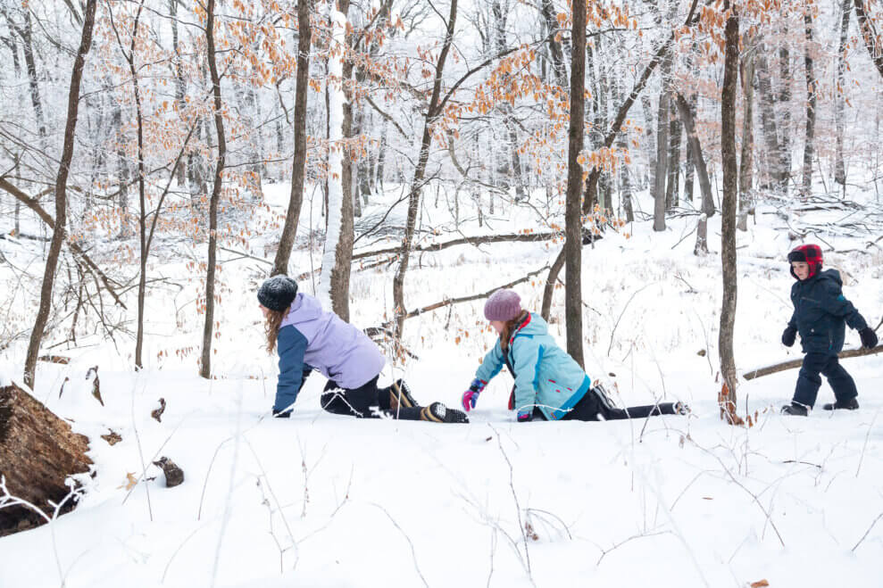 Two girls crawl through the snow in a forest, while their younger brother walks behind them on foot. 