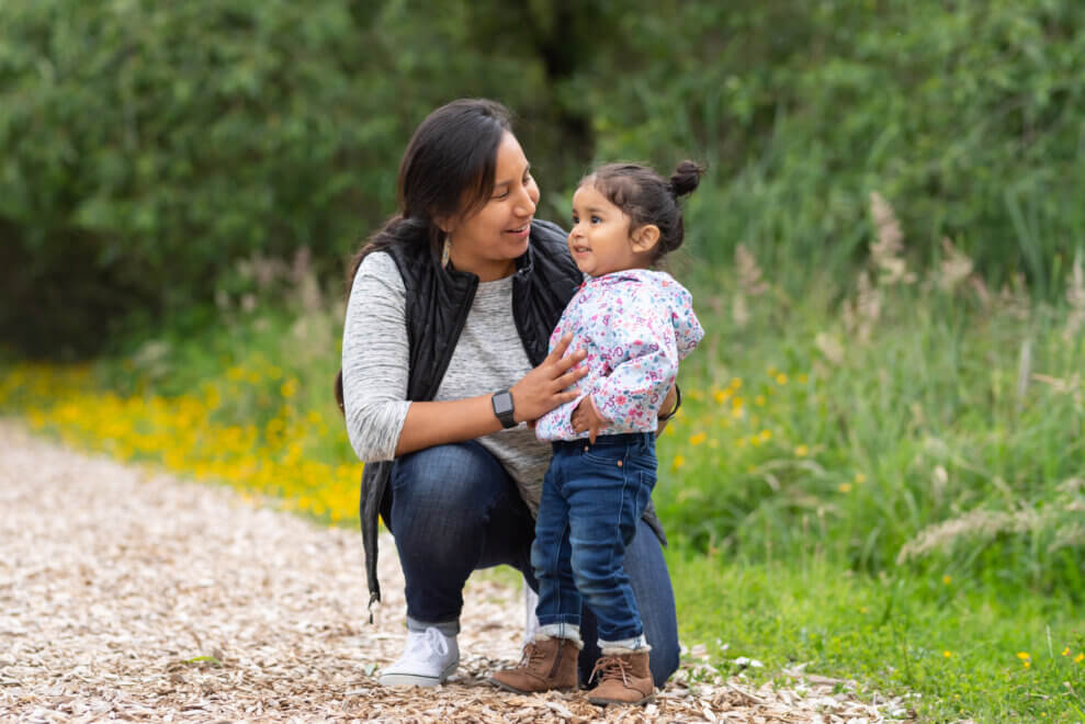 A mother crouches next to her toddler daughter on a trail and smiles at her.