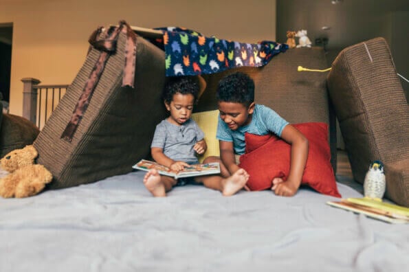 Two siblings sit inside a fort in their living room and read a book together.