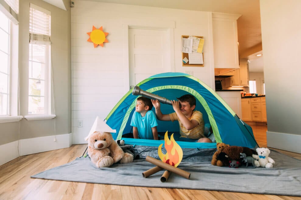 Two brothers sit in a blue tent in their living room. A fire cut out of construction paper rests on top of paper towel rolls that look like logs.