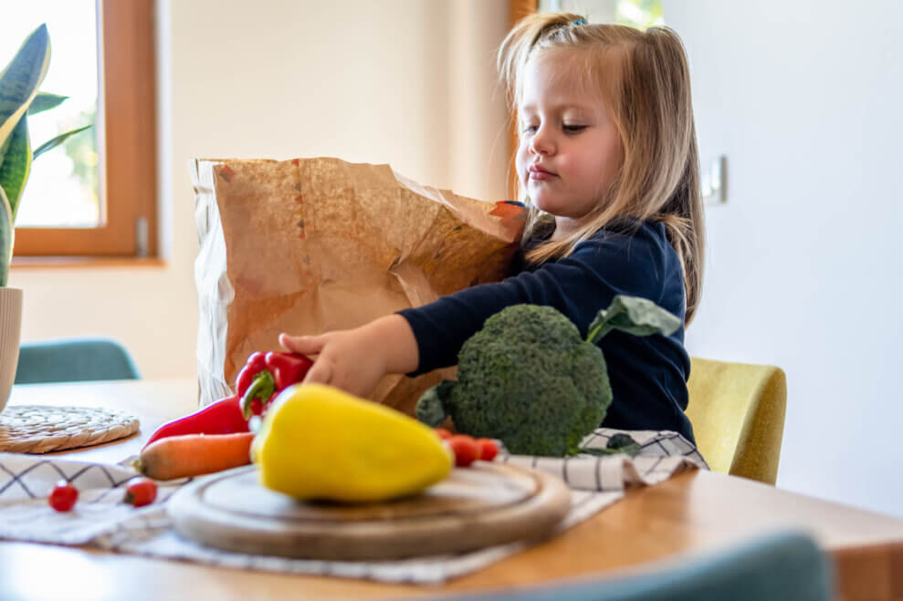 A young girl sits in a chair at the kitchen table and takes vegetables out of a brown paper grocery bag.