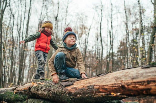 A boy crouches on a fallen log in the woods while his sister stands behind him, balancing with her arms out.