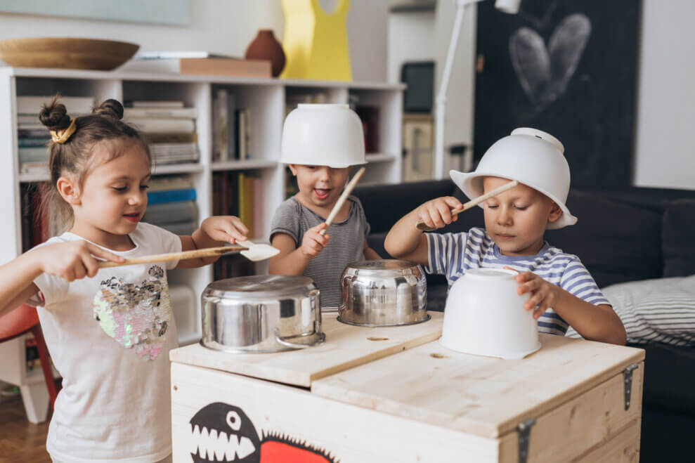 Two brothers and their sister stand in the living room, banging on pots and pants with kitchen utensils.