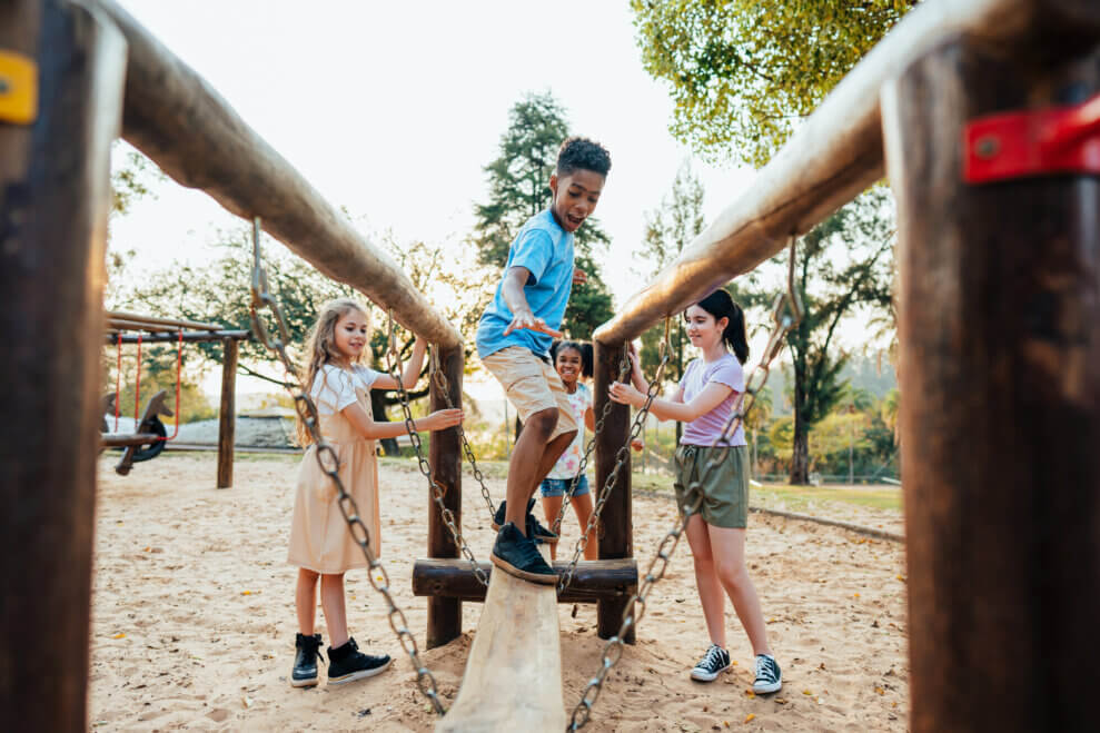 A group of four kids play on a wooden wobbly balance beam structure in an outdoor playground.
