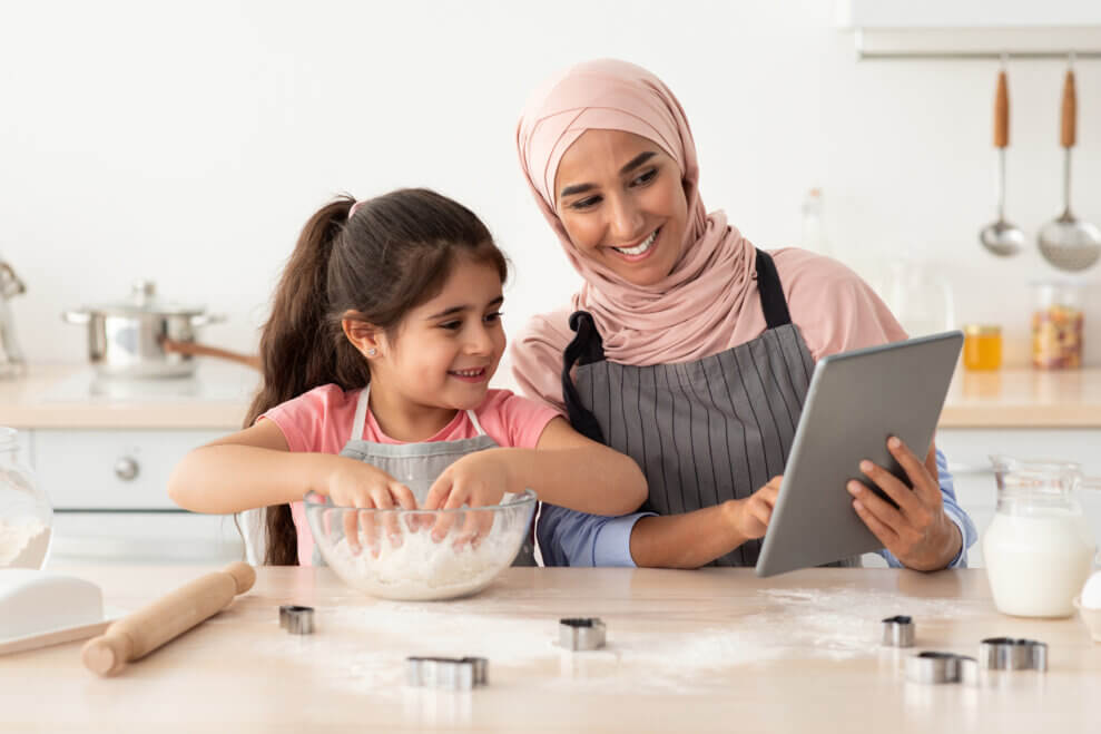 A mother and her daughter stand at the kitchen counter, baking together. The daughter has her hands in a bowl of ingredients, while her mother looks at a recipe on the iPad she's holding.