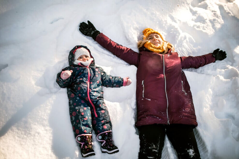 A mom and her two-year-old lie in the snow side-by-side and make snow angels. 