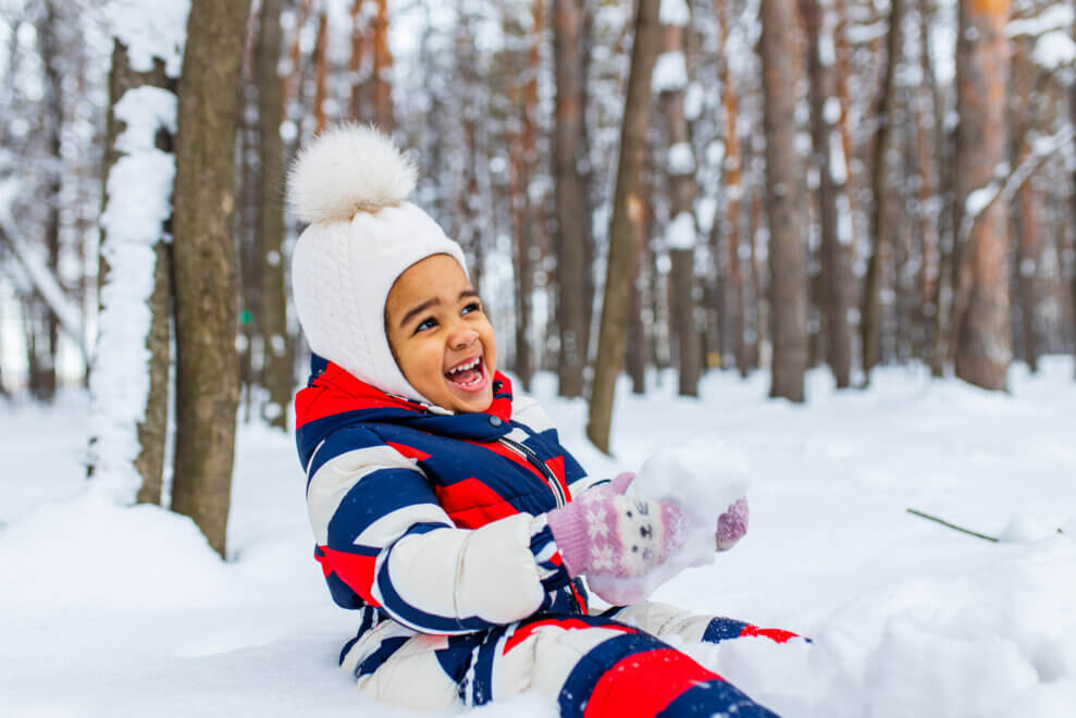A two-year-old girl sits in the snow, wearing a snowsuit and toque. She is smiling.