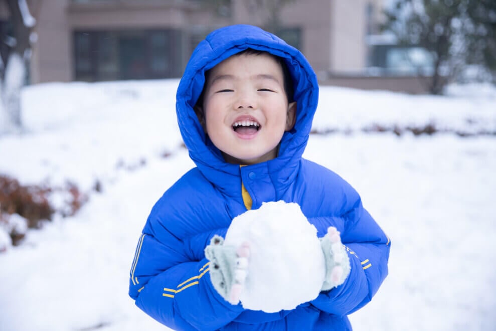 A toddler wearing a winter coat stands outside and holds a snowball in his mittened hands. He has a big smile on his face.