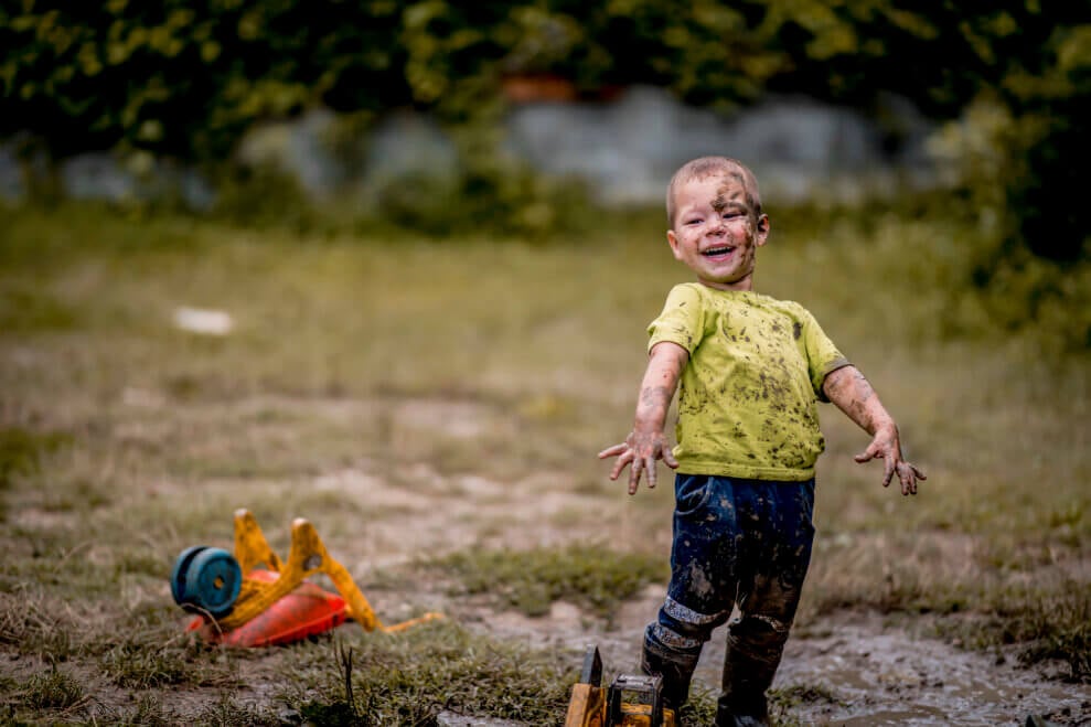 A young boy stands in a muddy field with a smile on his face. His clothes, boots, and face are covered in mud.