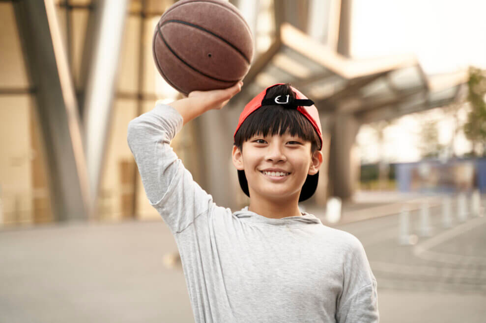 A smiling teenage boy holds a basketball over his head with one hand.