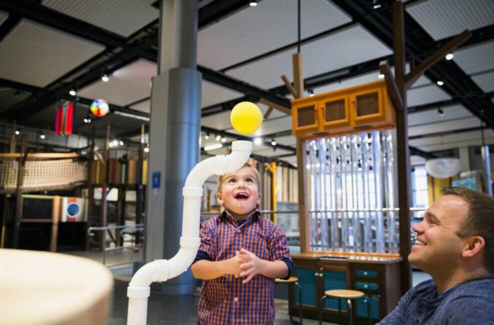 A boy and his father smile in awe at a yellow ball suspended above a pneumatic tube at the Creative Kids Museum.