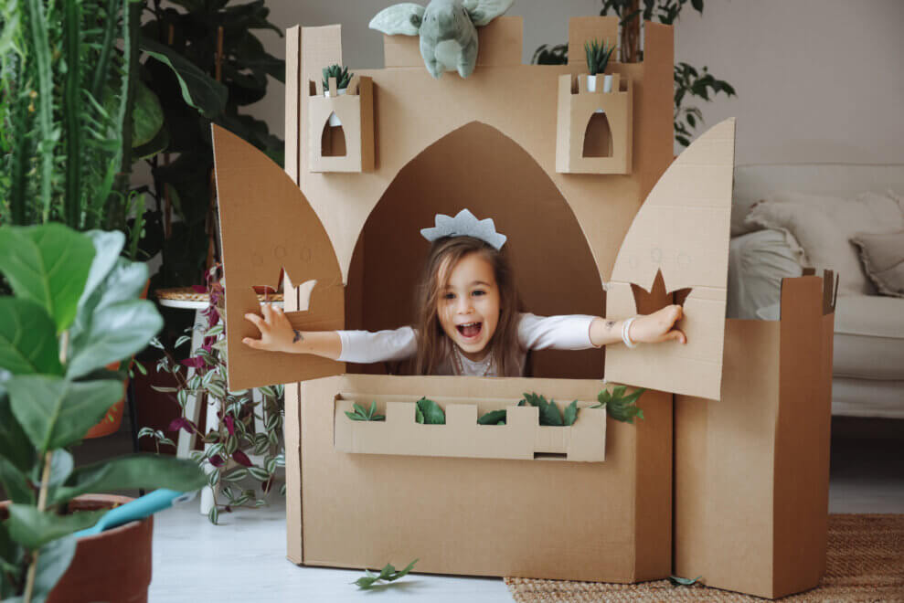 A little girl wearing a crown throws open the cut-out windows of her cardboard castle. She has a big smile on her face.