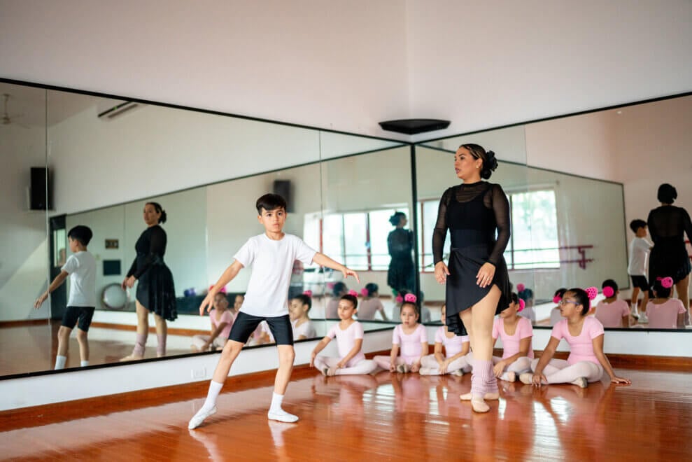 In a ballet class, a boy practices in front of his instructor while the girls in the class sit on the floor in front of the mirror.