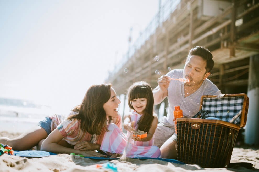 A family sits on a blanket on the each. The daughter plays with a sand toy while her father blows bubbles. 