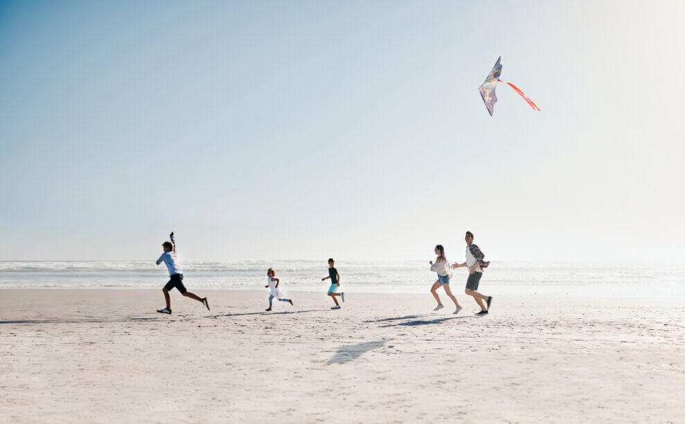 Three adults and two kids run across the beach as one of the adults flies a kite above them.