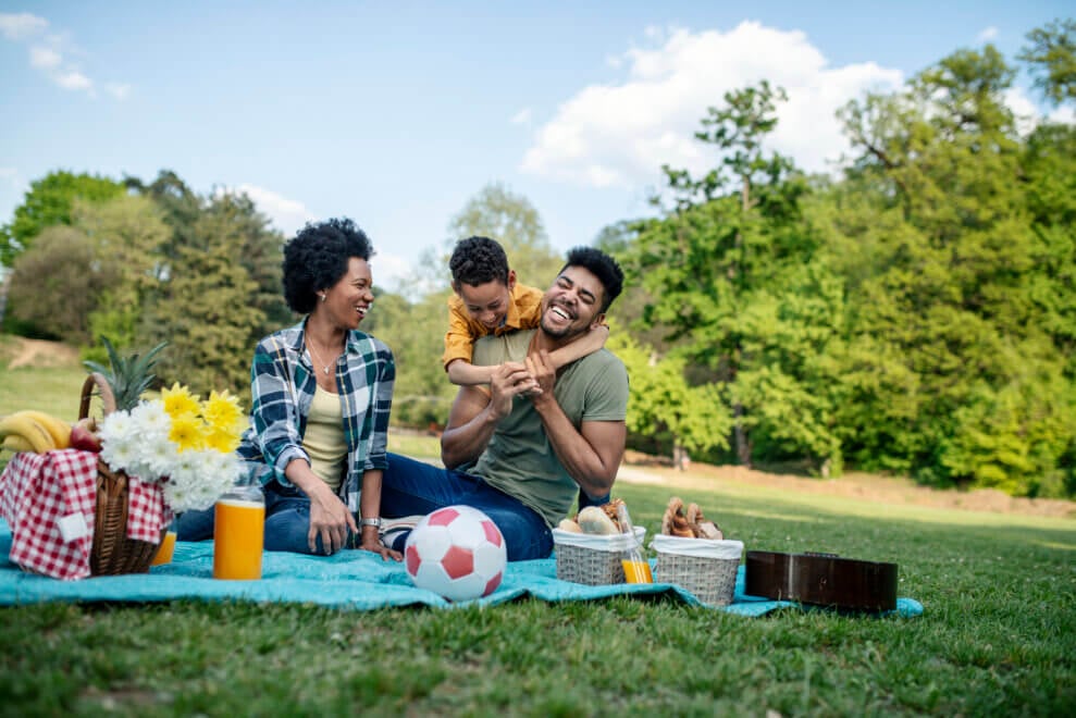 A family sits on a blanket together and has a picnic at a park. The son hangs off his father's shoulders as the mother and father laugh.