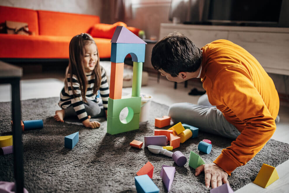 A father and his three-year-old daughter build a tower of blocks in the living room.