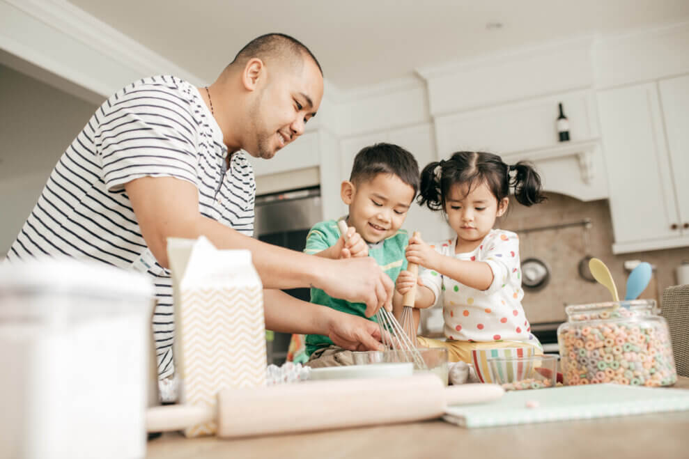 A father and his toddler son and daughter stand at the kitchen counter and mix baking ingredients in bowls.