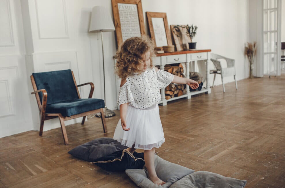 A three-year-old girl walks barefoot along a row of pillows lying on the floor in her living room.