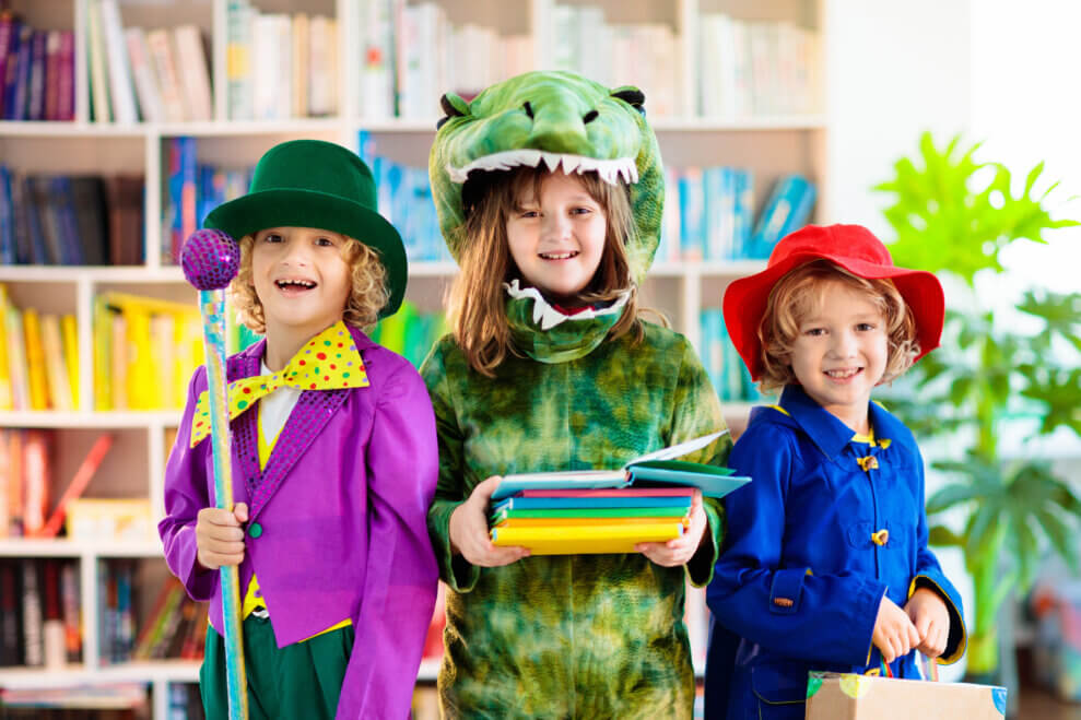 Three siblings stand in their living room, dressed up in costumes. One girl is a green dragon, another is a magician, and the the third is dressed like Paddington Bear.