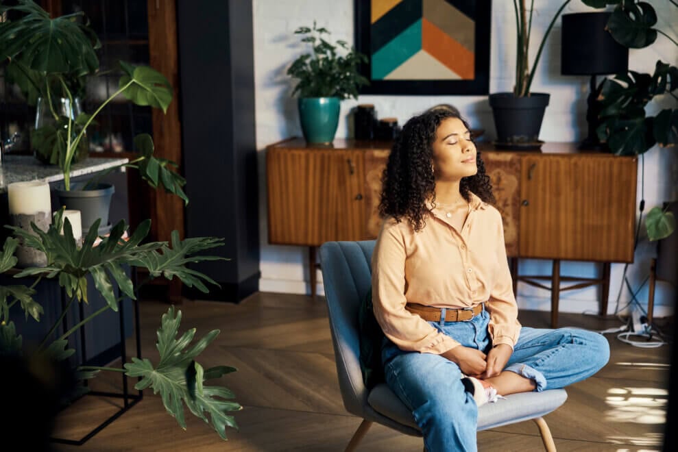 A woman sits on a chair in her living room, with her eyes closed and her hands in her lap. Light from the window shines on her face.