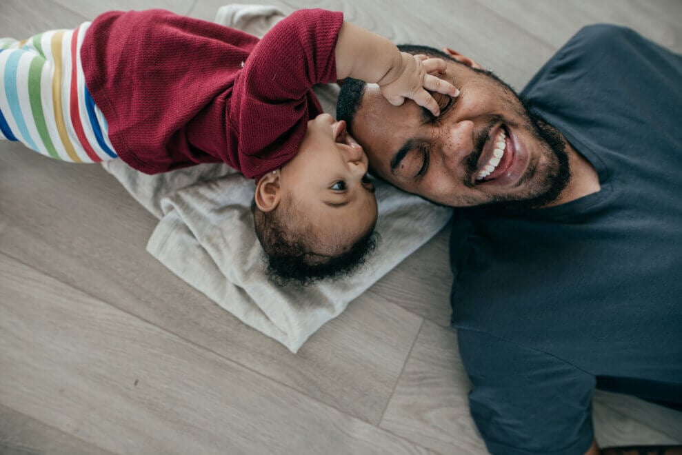 A father lies on his back on the floor next to his baby daughter. She reaches over to touch his face and he smiles.