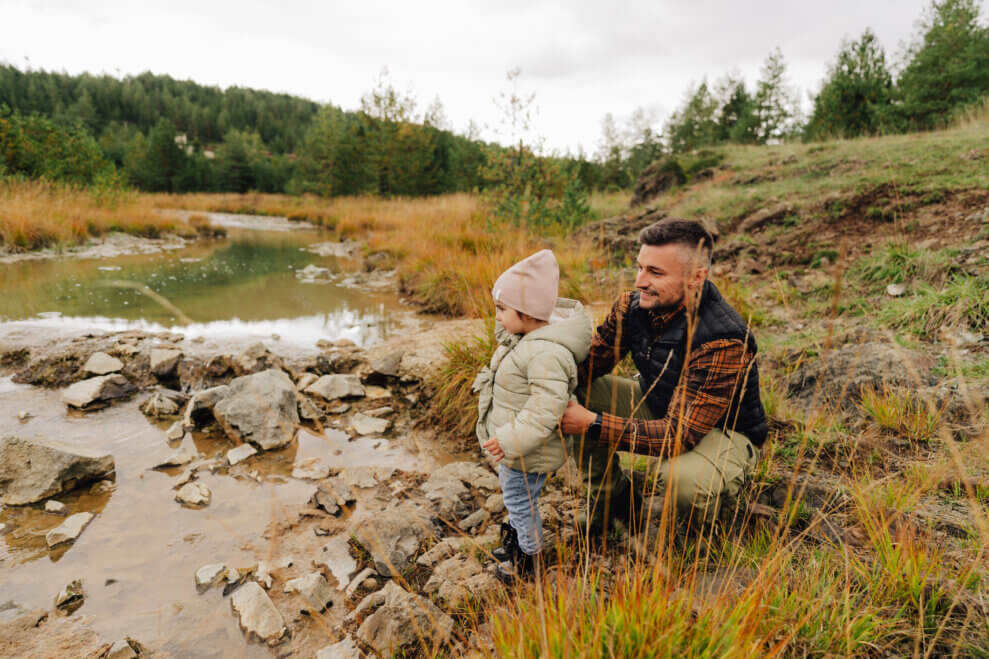 A father crouches behind his toddler daughter as she stands at the edge of a rocky creek bed.