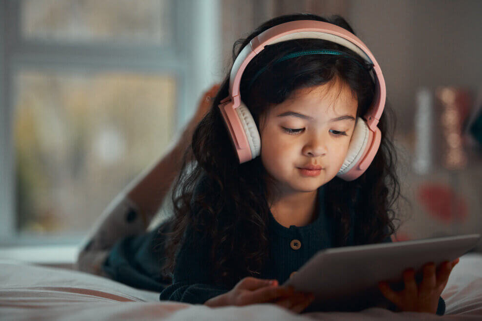 A young girl lies on her stomach, watching a show on an iPad. She wears pink headphones.