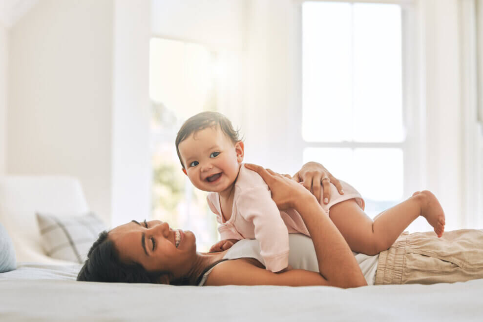 A mother lies on her back on her bed with her smiling baby resting on her stomach.