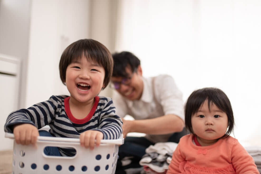 A grinning toddler sits in a laundry basket with his baby sister sitting beside him. Their father is behind them, folding laundry.